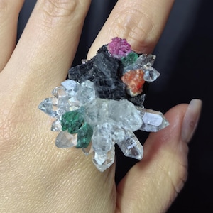 Könnte beinhalten: Ein einzigartiger Ring mit einem Cluster aus klaren Quarzkristallen, akzentuiert mit grünen, roten und schwarzen Mineralformationen. Das Design des Rings zeigt eine natürliche, organische Ästhetik, ideal für Liebhaber von Schmuck.