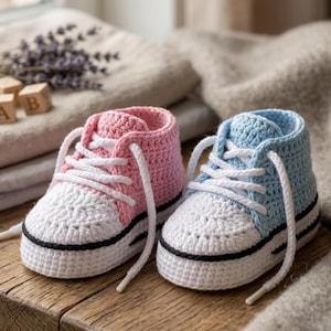 May include: A pair of handmade crochet baby sneakers. One is pink, the other is light blue, both with white toes, white laces, and black trim. The shoes are displayed on a wooden surface.