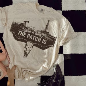 Op de afbeelding: Crèmekleurig T-shirt met een bruine afbeelding van olieboren, een schedel en de tekst "HOME IS WHERE THE PATCH IS". Het shirt ligt op een zwart-wit geblokt tapijt.