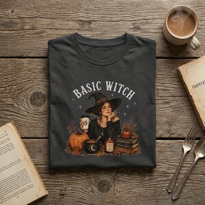 Könnte beinhalten: Ein dunkelgraues T-Shirt mit einer Grafik einer Person mit Hexenhut, die eine Kaffeetasse hält, umgeben von einem Kürbis, einem Kessel, einer Kerze und einem Stapel Bücher. Der Text "BASIC WITCH" ist in weißer Schrift darüber gedruckt.