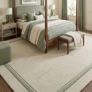 May include: A large rectangular area rug featuring a light beige center with a sage green border and double-line detailing. The rug is positioned in a bedroom setting beneath a wooden canopy bed, matching the sage green bedding and wall accents.