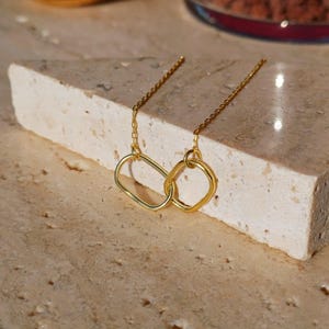 May include: Gold-colored earrings featuring two linked geometric shapes: an oval and a square. The earrings hang from delicate gold chains. The jewelry is displayed on a light-colored, textured surface.