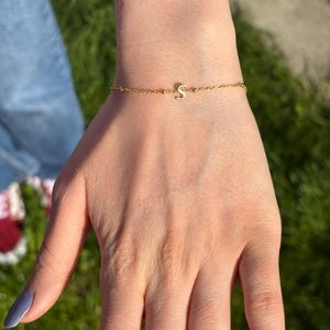 Op de afbeelding: Een delicate goudkleurige armband met een kleine letter "S" bedel. De armband is een fijne ketting met kleine kraaltjes. De armband zit om een pols met een blauw gelakte nagel.
