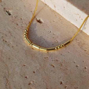 Puede incluir: Un delicado collar de oro con una serie de pequeñas cuentas cilíndricas y esféricas. El collar se muestra sobre una superficie clara y texturizada, creando un contraste con el oro brillante. La cadena es fina y flexible.