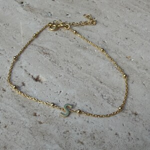 Op de afbeelding: Een delicate goudkleurige armband met de letter "S" als centrale bedel. De armband bestaat uit een fijne ketting afgewisseld met kleine, ronde kralen. De verstelbare sluiting zorgt voor een comfortabele pasvorm. Ongeveer 15 cm lang.
