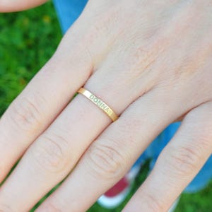 Pode incluir: Um anel dourado com o nome "DONNA" gravado na banda. O anel está num dedo com verniz lilás claro. O fundo inclui relva verde e calças de ganga azuis.