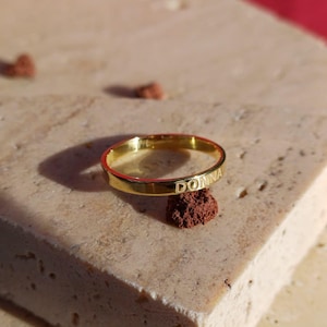 Peut inclure: Une bague dorée avec le nom "DONNA" gravé. La bague est posée sur une surface claire et texturée. La bague est de conception simple, avec les lettres du nom clairement visibles. C'est un bijou.