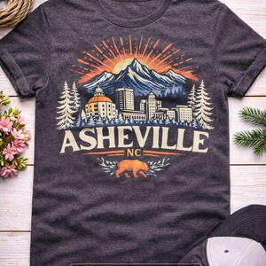 May include: Dark heather gray t-shirt featuring a mountain and city landscape design with the text "Asheville NC". The design includes a bear graphic and a rising sun. A baseball cap is visible in the image.