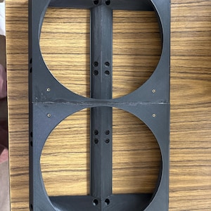 May include: A black, rectangular, 3D-printed object with two large circular openings. The object has a central vertical support and is secured with small brass-colored screws. The item is likely a component or housing.
