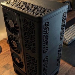 May include: Black computer case with geometric cutouts and three fans visible on the side. The case has a textured finish and a patterned top. The design incorporates ventilation for optimal performance. The case is on a wooden surface.