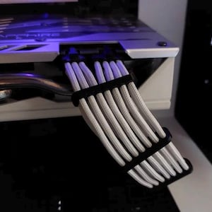 May include: Close-up of a computer's internal components, showcasing white braided cables neatly organized with black cable combs. The cables are connected to a silver and white component, with a glimpse of blue lighting.