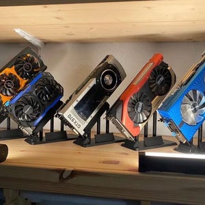 May include: A collection of five colorful graphics cards, each mounted on a black stand. The cards feature various designs with orange, blue, red, and silver accents. The text "GTX 970" is visible on one card. The cards are displayed on a wooden shelf.