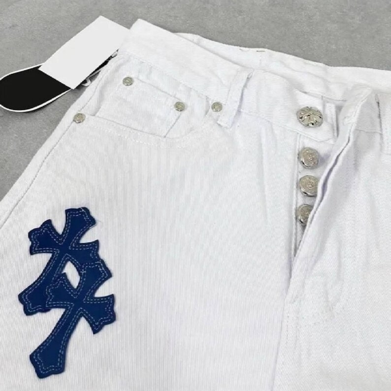 Peut inclure: Jean blanc en denim avec fermeture &agrave; boutons et rivets argent&eacute;s. Un &eacute;cusson bleu fonc&eacute; en forme de croix est cousu sur le devant. Une &eacute;tiquette blanche avec une &eacute;tiquette noire est attach&eacute;e au jean.