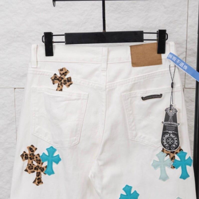 May include: White denim jeans with decorative cross patches in leopard print and turquoise. The jeans have a classic five-pocket design and a leather patch on the back. The jeans are hanging on a black hanger.