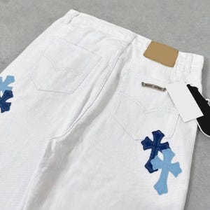 Peut inclure: Jean blanc avec deux poches arri&egrave;re et un petit patch marron. Le jean pr&eacute;sente deux &eacute;cussons en forme de croix dans les tons de bleu sur les poches arri&egrave;re. Une petite &eacute;tiquette m&eacute;tallique est visible sur la poche arri&egrave;re.