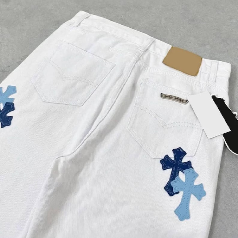 Peut inclure: Jean blanc avec deux poches arri&egrave;re et un petit patch marron. Le jean pr&eacute;sente deux &eacute;cussons en forme de croix dans les tons de bleu sur les poches arri&egrave;re. Une petite &eacute;tiquette m&eacute;tallique est visible sur la poche arri&egrave;re.