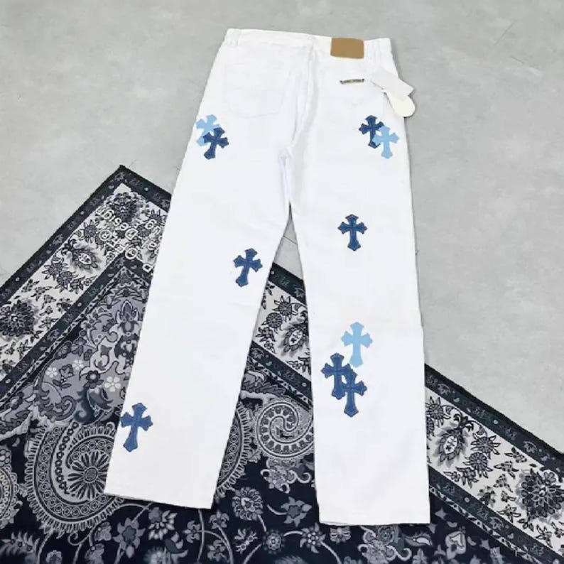 Peut inclure: Jean blanc avec plusieurs appliques de croix bleues. Le jean a une conception classique &agrave; cinq poches et une coupe droite. Un patch en cuir marron est visible sur la ceinture arri&egrave;re.