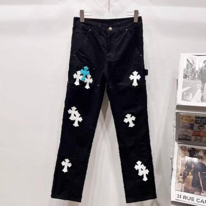 May include: Black corduroy pants with white cross appliques. One cross is light blue. The pants have a classic five-pocket design and a button and zipper closure. The pants are hanging on a hanger.