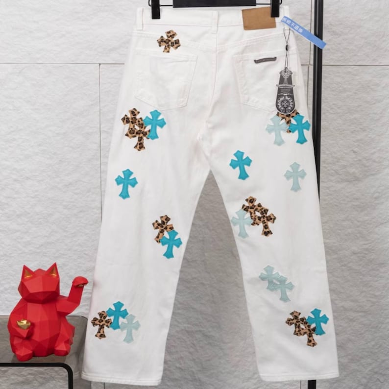 May include: White denim jeans with multiple cross designs. The crosses are in various styles, including solid turquoise, leopard print, and a combination of both. The jeans have a classic five-pocket design.