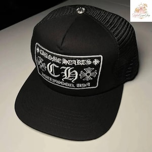 Puede incluir: Gorra de camionero negra con parte trasera de malla y visera curva. La parte delantera presenta un parche bordado blanco con el texto "CHROME HEARTS" y "MIDWOOD USA". Un emblema plateado está en la parte superior.