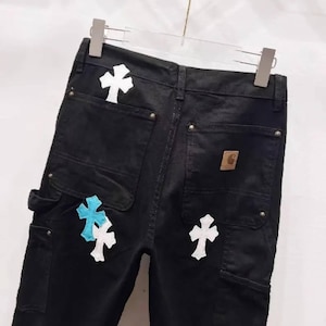 May include: Black denim work pants with white and light blue cross patches. The pants have multiple pockets, a brown Carhartt patch, and are hanging on a metal hanger.