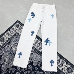 Puede incluir: Vaqueros blancos con múltiples aplicaciones de cruces azules. Los vaqueros tienen bragueta con botones y un diseño clásico de cinco bolsillos. Las cruces son de diferentes tonos de azul.