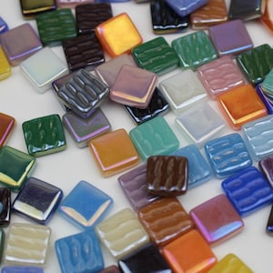 May include: A collection of small, square mosaic tiles in various colors, including blue, green, orange, and purple. Some tiles have a textured surface, while others are smooth. The tiles are scattered on a white surface.