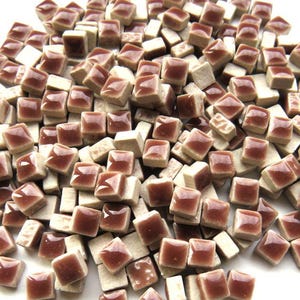 May include: A pile of small, square ceramic tiles. The tiles have a glossy, reddish-brown surface and are interspersed with off-white tiles. The tiles are scattered on a white surface.
