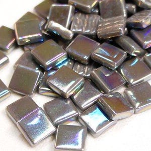 May include: A pile of iridescent square tiles. The tiles have a metallic sheen with colors shifting from silver to purple and blue. The tiles are small, with a slightly rounded top and a square shape. They are suitable for mosaic projects.