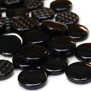 May include: A collection of small, round, black glass tiles. Some of the tiles have a smooth, glossy surface, while others feature a textured, grid-like pattern on their top side. The tiles are scattered in a pile, showing their dark color.