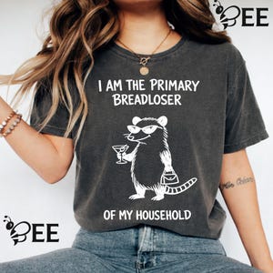 Könnte beinhalten: Ein anthrazitfarbenes Kurzarm-T-Shirt mit einer weißen Grafik eines Opossums, das eine Sonnenbrille trägt und ein Martiniglas sowie eine Handtasche hält. Der Text lautet I AM THE PRIMARY BREADLOSER OF MY HOUSEHOLD in weißen Buchstaben.