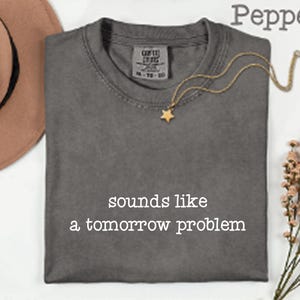 Peut inclure: Un t-shirt à col rond gris anthracite avec le texte blanc "sounds like a tomorrow problem" imprimé sur le devant. Le t-shirt présente une étiquette de marque visible à l'intérieur du col et un design à manches courtes en tissu uni.