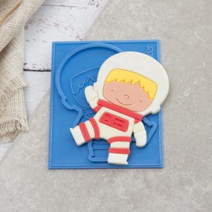 May include: A white, red, and yellow astronaut-shaped cookie cutter. The astronaut has a white helmet, suit with red accents, and yellow hair. The cookie cutter is on a blue rectangular base.