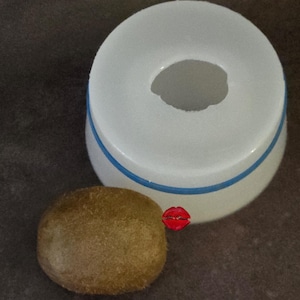 May include: A white, circular kitchen tool with a central opening and a thin blue horizontal stripe, positioned next to a single brown, fuzzy kiwi fruit. A small red logo with the text Lillie Classic is visible on the side of the white tool.