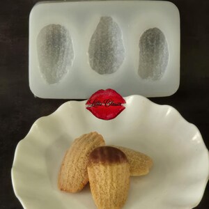 May include: A rectangular translucent silicone mold with three shell-shaped cavities positioned above a white scalloped bowl containing three golden-brown madeleine cakes, one of which is partially dipped in dark chocolate, with a red lip logo.
