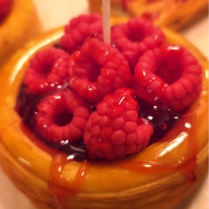 May include: A round pastry topped with a cluster of bright red raspberries and a dark purple fruit filling. A thin white candle wick protrudes from the center of the fruit. The pastry has a golden-brown crust with a glossy, translucent red glaze.