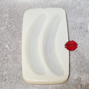 May include: A clear, rectangular silicone mold with two banana shapes. The mold has a smooth surface and is set against a gray background. A red lip-shaped sticker with the text "Little Classic" is on the right side.
