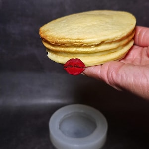 May include: A stack of four golden-brown, round objects, possibly soap or resin, with a gold shimmer on the edges. A red lip-shaped logo with the text "Little Classes" is visible. A clear, circular mold sits below.