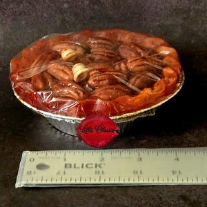 May include: A small pecan pie in a silver aluminum foil pan, wrapped in clear plastic film. A white ruler is positioned in front, showing the pie has a diameter of approximately 4 inches. A red lip-shaped logo with the text Lilliz Classix is visible.