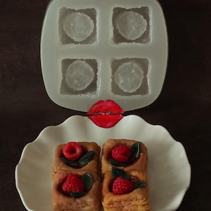 May include: A translucent silicone mold with four square cavities and a white scalloped dish containing four square pastry-like items topped with red raspberries and dark green leaves. A red lip-shaped logo with the text Lillie Classic is visible.