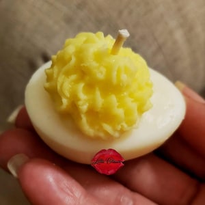May include: A deviled egg shaped candle with a smooth white base and a textured, bright yellow piped center. A small white wick protrudes from the top of the yellow portion. A red logo with the text Lillie Classix is visible on the white base.