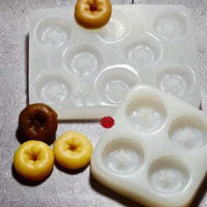 May include: Two translucent silicone molds for miniature ring-shaped pastries, one rectangular with eight cavities and one square with four cavities. Three small, round, golden-brown and yellow donut-shaped items are placed nearby.