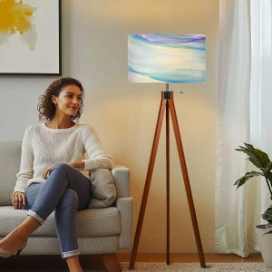 May include: A floor lamp with a three-legged wooden tripod base and a rectangular fabric lampshade. The lampshade features a soft, abstract watercolor pattern in shades of blue, purple, and white, emitting a warm, diffused light.