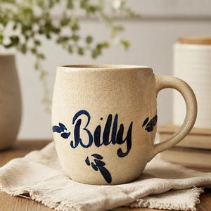 May include: A beige ceramic mug with a curved handle, featuring the name "Billy" in dark blue script. The mug sits on a beige linen napkin, with a vase and a container with a wooden lid in the background.