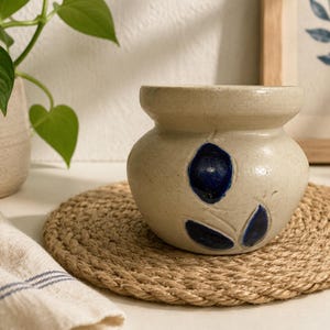 May include: A beige ceramic pot with dark blue floral designs, sitting on a round, woven jute coaster. A potted plant and a folded linen towel are in the background. The pot has a rounded body and a flared rim.