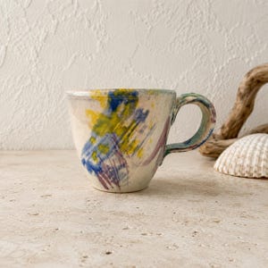 May include: A ceramic mug with a cream-colored base featuring abstract brushstrokes in shades of blue, yellow, and purple. The mug has a rounded body and a curved handle, with the painted design extending onto the handle surface.