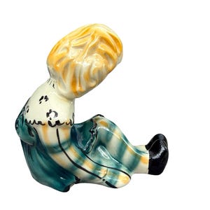 May include: A vintage ceramic figurine of a seated figure with blonde hair, wearing a teal and yellow plaid dress. The figurine has black shoes and a white collar with black floral accents. The figure is in a seated position.