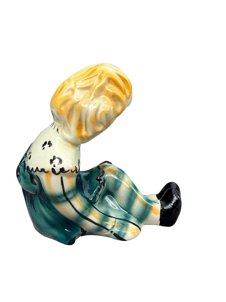 May include: A vintage ceramic figurine of a seated figure with blonde hair, wearing a teal and yellow plaid dress. The figurine has black shoes and a white collar with black floral accents. The figure is in a seated position.