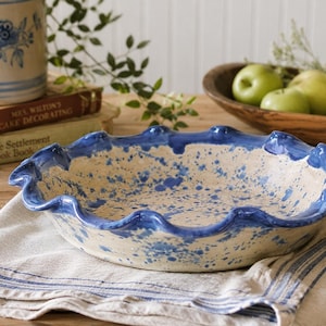 May include: A ceramic pie dish with a wavy, blue-painted rim and a speckled interior. The dish is set on a striped linen cloth, with a wooden bowl of green apples in the background. A stoneware jar and cookbooks are also visible.