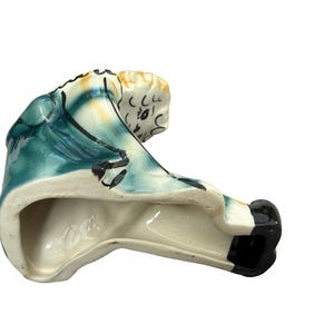 May include: A ceramic figurine of a seated figure, painted with teal, cream, and black accents. The figure has a detailed face and is wearing a teal garment. The base is unglazed, revealing the ceramic material.
